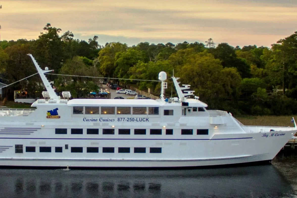An exterior shot showcasing a large white yacht with casino signage, surrounded by trees and waterfront views. The scene conveys leisure, excitement, and the possibility of a unique on-the-water experience.