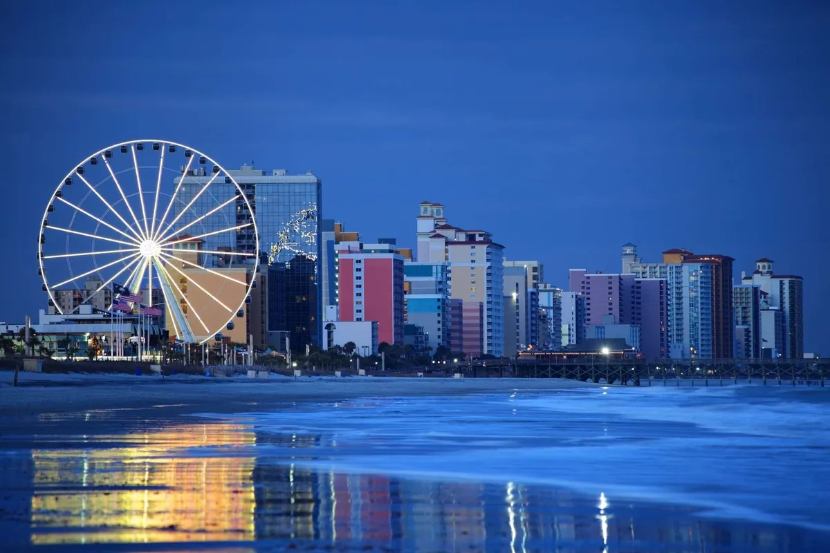 A vibrant scene featuring a Ferris wheel, festive beachfront buildings, and rolling ocean waves. The area is lively and colorful, suggesting nearby attractions and opportunities for evening exploration and upscale dining.