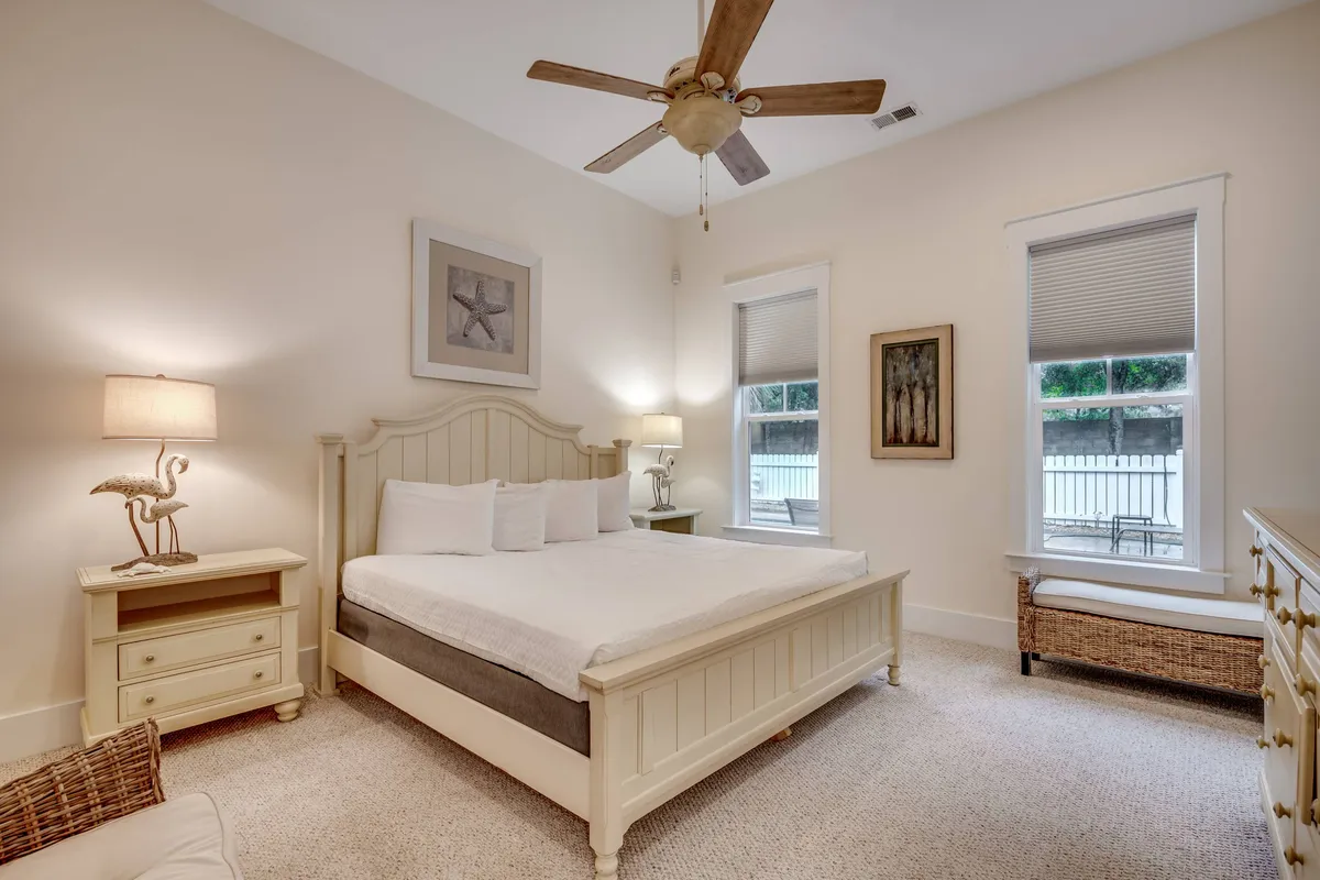 A serene bedroom featuring a plush bed, soft bedding, ceiling fan, bedside lamp, and tranquil artwork, bathed in natural light from large windows. The inviting atmosphere is ideal for restful nights or leisurely mornings.