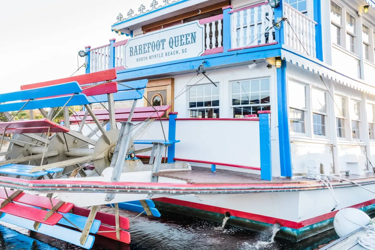 The colorful paddlewheel boat 'Barefoot Queen' docked at the waterfront, with its eye-catching sign and lively facade. The scene is bustling with anticipation for a sightseeing or dinner cruise, linking to the complimentary family activity package mentioned, including the Barefoot Queen Riverboat Dinner Cruise.