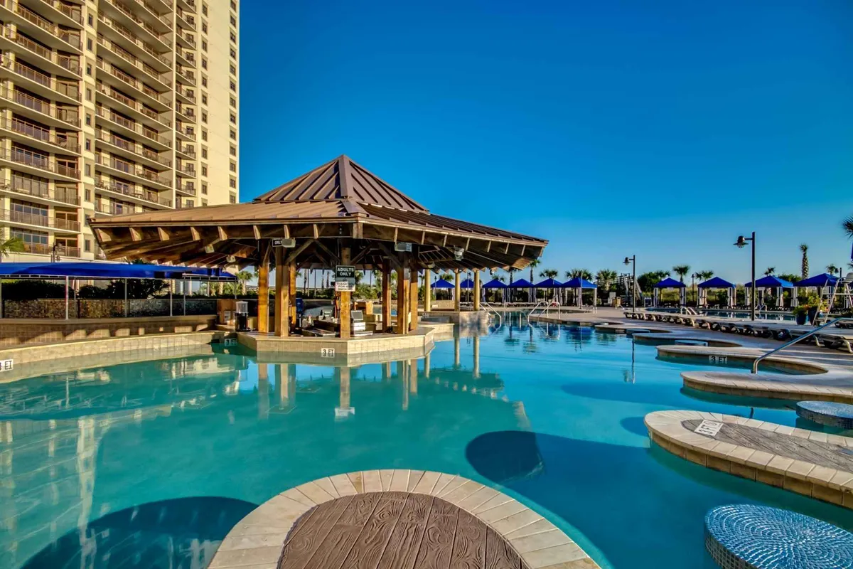 A vibrant resort pool complex with a large swimming pool, poolside cabana, and plush lounge chairs shaded by blue umbrellas. The setting is lively and inviting, suggesting ample space for families to play, swim, and unwind together, echoing the mention of a sprawling pool complex and shared resort amenities.