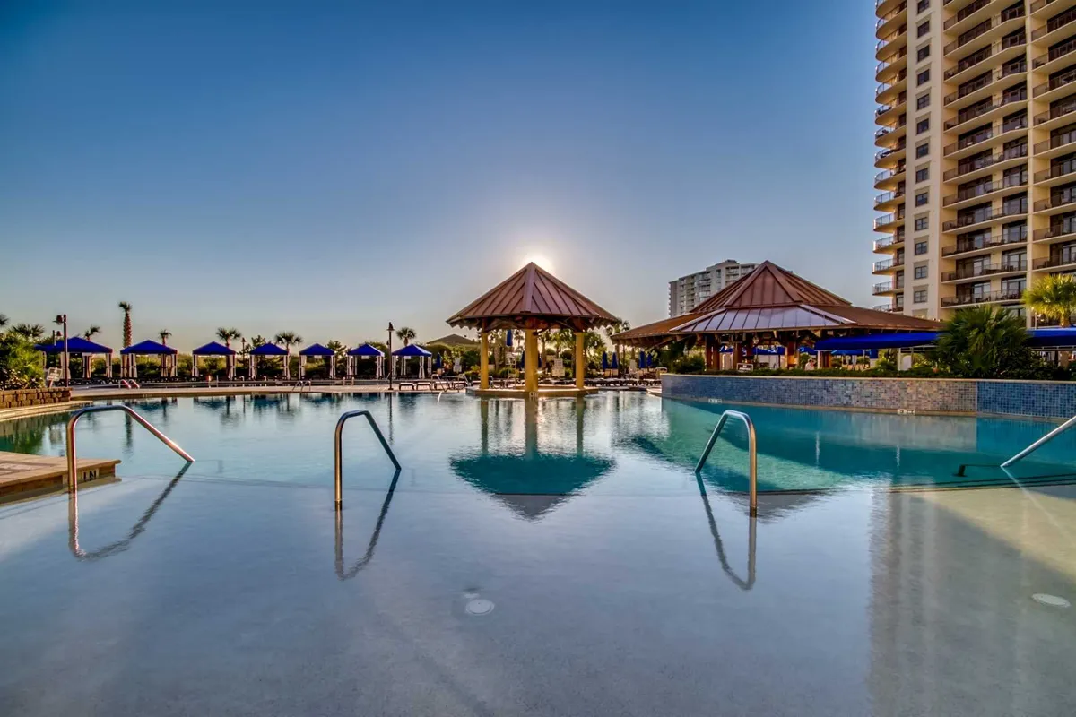 A serene resort swimming pool is surrounded by tropical cabanas and palm trees, with the sun setting in the background. The tranquil water, inviting lounge chairs, and luxurious atmosphere evoke relaxation and high-end resort amenities.