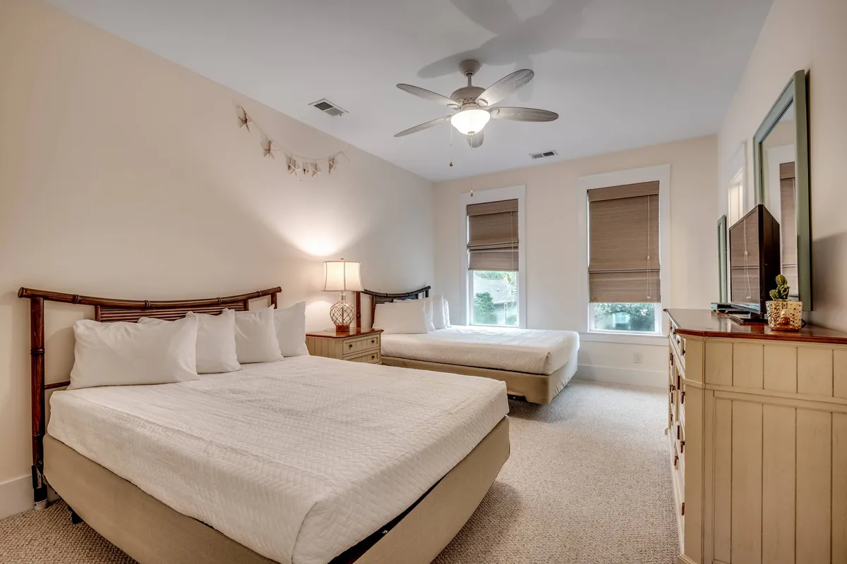 A welcoming bedroom with two beds, a nightstand with a lamp, a dresser, and windows that allow natural light to fill the space. The layout suggests room for multiple guests, ideal for families or groups.