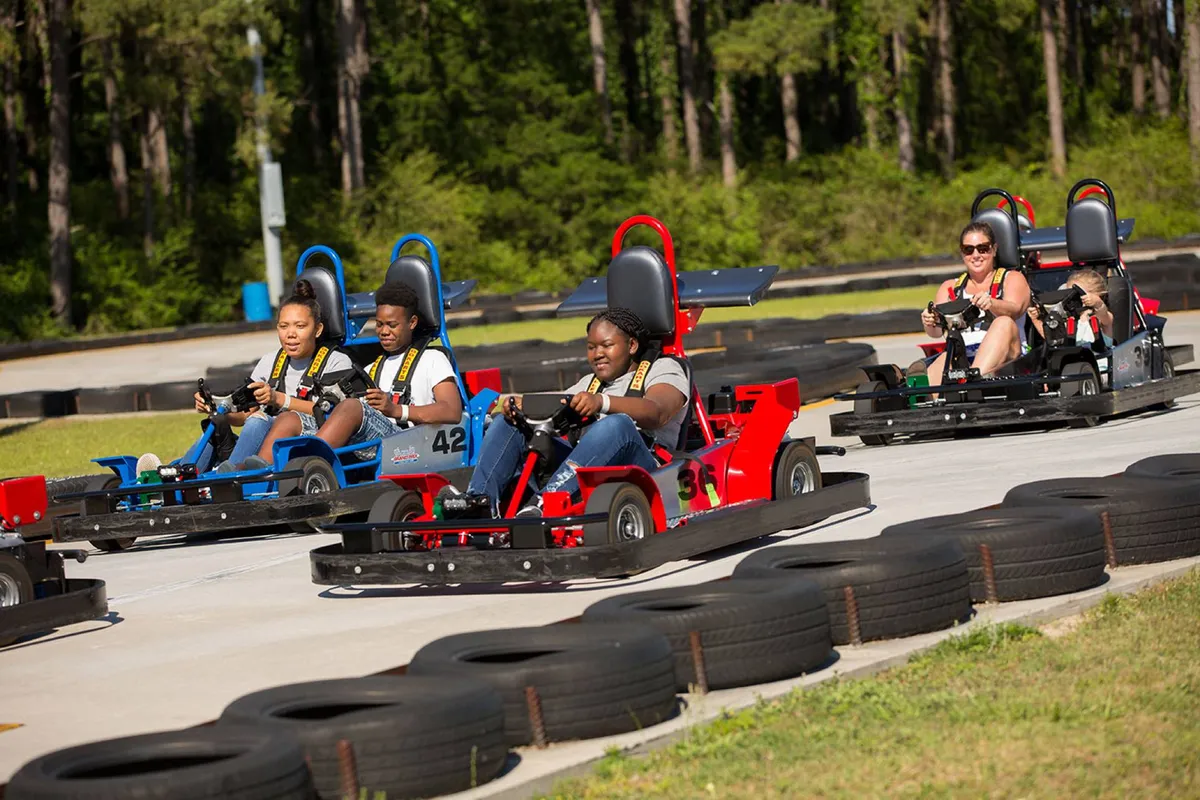 An action-packed scene at an outdoor go-kart track, with participants racing around curves amid safety barriers and a lively atmosphere. The image conveys a sense of playful competition and adventure, matching the thrill-seeker attractions mentioned in the text.