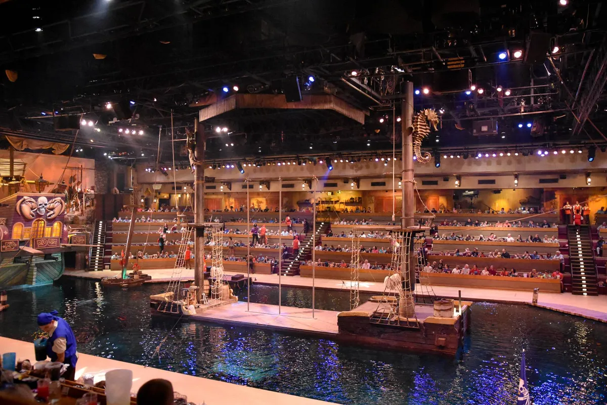 A vibrant shot of a themed theater stage, complete with water features and decorative ship elements, viewed from the audience seating area. The atmosphere is festive, hinting at immersive live performances and group entertainment.